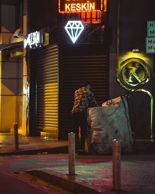 A person in a camouflage-patterned jacket is standing on the pavement outside a commercial building at night, handling a large, irregularly shaped bag or sack made of heavy, weather-resistant fabric. The scene is illuminated by artificial lighting, including neon signs and streetlights, casting a glow on the surrounding area. The building features a black roller shutter door, partially closed, with a bright, diamond-shaped illuminated sign above it and a neon sign displaying the word 'KESKIN' in red above that. To the right of the person, there is a circular, metallic object attached to the building's exterior wall, possibly an decorative element or utility fixture, with a small green light or sign nearby. In the foreground, two short bollards made of metal or plastic, painted with reflective stripes, separate the sidewalk from the road. The environment appears urban, with elements typical of city streets at night, reflecting a scene where private waste handling or rubbish collection might be occurring outside a commercial premise, supported by the presence of rubbish bags and the setting within a busy street area.