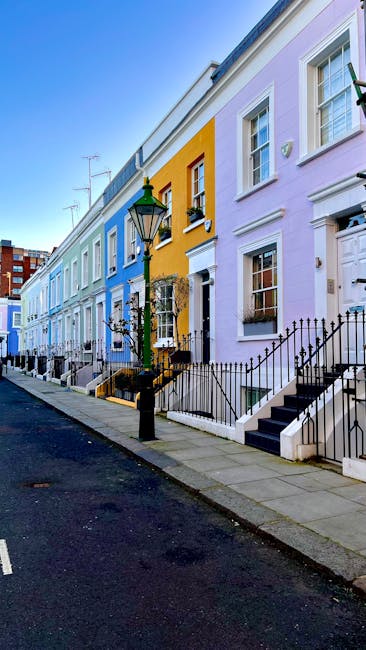 A row of colorful terraced houses situated along a narrow urban street, with each building painted in pastel shades including light blue, yellow, and lavender. The houses feature white-framed sash windows and decorative moldings, with some including small front gardens enclosed by black wrought iron railings. A traditional black street lamp stands in the foreground, illuminating the pavement and steps leading up to the entrances. The pavement is made of grey concrete slabs, with a dark asphalt road running along the street. The sky is clear and blue, suggesting daytime weather. The scene appears well-maintained and typical of residential neighborhood areas where private waste collection services, like those provided by House Clearance West Kensington, might manage rubbish removal for individual properties or street waste clearance, supporting alternative waste handling methods outside local authority trash collection.