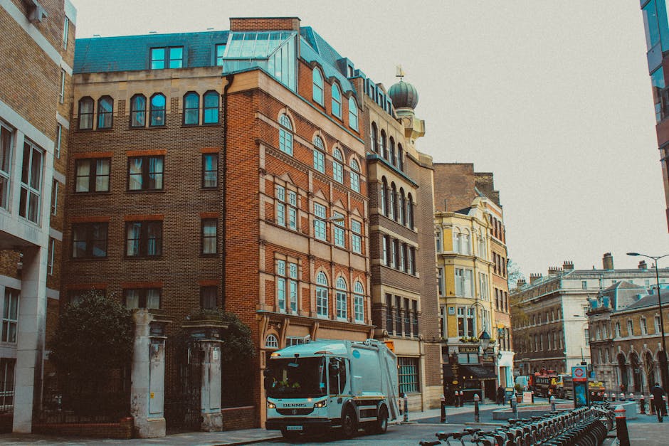 A city street scene featuring a white waste collection vehicle parked on the right side of the road, partially blocking the pavement. The vehicle is positioned near a row of dark metal bike racks and is close to a gated entrance with stone pillars, leading to a residential property. Surrounding the vehicle are tall, multi-storey buildings showcasing a mixture of brickwork and ornate architectural details, including arched windows and decorative cornices. The buildings display a range of colors, from reddish-brown bricks to cream and pale yellow facades, with some having metal roofs or domed roofs with finials. The scene is evenly lit with natural daylight, and the background reveals more historic structures with intricate facades lining the street, which is quiet with a few pedestrians and parked bicycles. The presence of the waste collection vehicle from HouseClearanceWestKensington.co.uk relates to private waste handling and alternative rubbish removal options in this urban environment.