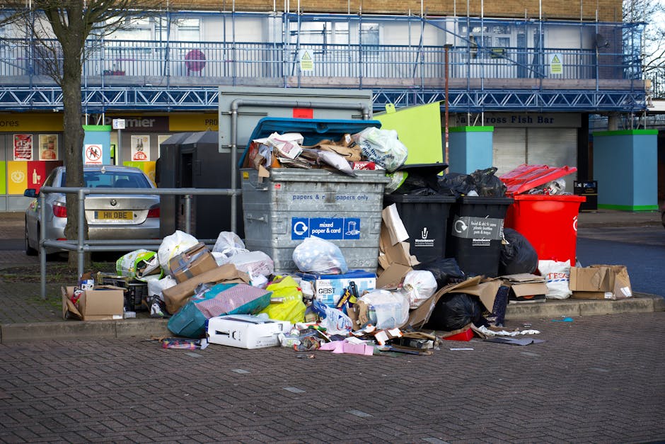 A large collection of mixed household waste and rubbish, including cardboard boxes, plastic bags, paper parcels, and discarded packaging, is piled up on a paved parking area. The rubbish is situated in front of a set of black, green, and red wheelie bins, with one bin lid partially open revealing more trash inside. In the background, there are two cars parked behind a metal railing, with one silver vehicle visible. Further behind, a storefront with a yellow sign and large glass windows can be seen, along with a building wrapped in blue construction scaffolding and netting, indicating ongoing renovations. The scene is lit by natural daylight, with the waste materials sprawling across the pavement beneath a tall, leafless tree standing on the left side of the image. This setting illustrates typical urban rubbish accumulation outside a commercial or residential property, which private waste collection services such as those offered by houseclearancewestkensington.co.uk might manage as an alternative to official council rubbish collection, especially for household waste requiring disposal or clearance.