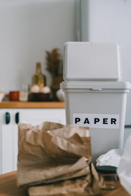 A white rectangular rubbish bin with a hinged lid, positioned upright on a kitchen countertop or table, with the word 'PAPER' in bold black letters on an adhesive label attached to its front. The bin is made of plastic with a smooth finish and is open, revealing an empty interior. In the foreground, there is a crumpled brown paper bag resting on the surface, along with some scattered white paper and other small waste items nearby. The background shows a blurred kitchen scene with a wooden shelf or countertop, some bottles, containers, and decorative objects, suggesting an indoor domestic environment. The lighting is natural and soft, highlighting the clean and organized theme consistent with waste management and rubbish removal practices, often associated with services provided by companies like houseclearancewestkensington.co.uk, especially when dealing with household waste and paper recycling.