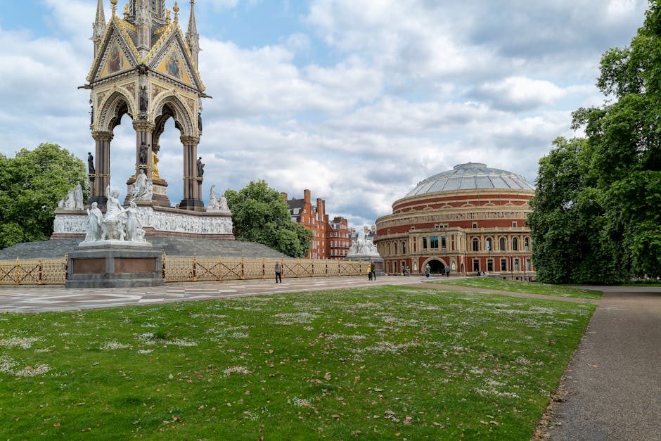 The image shows a large, open park area with a well-maintained grassy surface scattered with small white flowers, bordered on one side by a paved pathway. In the background, there is an ornate, multi-tiered monument featuring detailed architectural elements, sculptures, and decorative columns, predominantly made of stone with a combination of light and dark finishes, situated on a raised platform with a surrounding low fence. Behind the monument, there are trees with lush green foliage and a historic brick building with circular architecture and arched windows, which appears to be part of an indoor or exhibition venue. The scene is set during daytime under a partly cloudy sky, with diffused natural light illuminating the surroundings evenly. The area looks clean and free of debris, consistent with a maintained public space that might be relevant for private waste clearance or event-related rubbish removal services, such as those offered by houseclearancewestkensington.co.uk, operating in West Kensington. The scene emphasizes the importance of careful site management during large outdoor gatherings or events to ensure cleanliness and environmental quality.