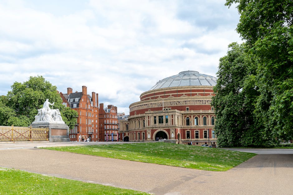 A wide daytime view of a historic, round concert hall building with a domed roof, constructed with red brick and ornate stone detailing around its windows and entrances, situated on a green lawn with a paved pathway leading towards it. To the left of the building, there is a white marble statue of multiple figures positioned on a stone pedestal, partially obscured by lush green trees that frame the scene on both sides. The sky is partly cloudy, casting soft, diffused light over the area. In the background, several red-brick residential buildings with pitched roofs and chimneys are visible behind the trees and the concert hall, creating an urban park atmosphere. The scene appears peaceful and well-maintained, with no visible debris or waste, reflecting the environment in which independent and private waste disposal services by companies such as houseclearancewestkensington.co.uk might operate to support clean, organized public spaces during events or general maintenance.