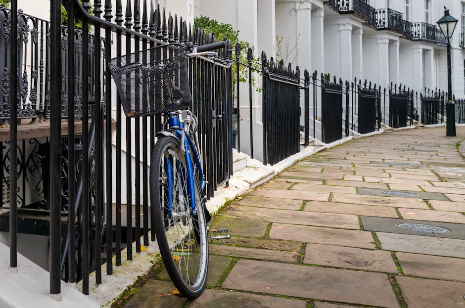 In the foreground, a blue bicycle with a black front basket leans against a tall black metal fence with pointed finials, which runs along the edge of a paved sidewalk. The bicycle appears to be a city bike with a textured, matte-finished frame, and silver components such as the spokes and pedals. Behind the fence, white terraced houses with black wrought iron balconies, decorative white columns, and traditional street lamps are visible, indicating a residential area. The pavement consists of large rectangular stone slabs, some with slight moss or dirt in the joints, curving gently to the right in the background. The scene appears to be captured in natural daylight with soft, even lighting, emphasizing the textures of the stone, metal, and painted surfaces. The overall setting suggests a typical urban street scene where private transport and street furniture coexist, aligning with the context of local garden, house, or rubbish clearance services provided by companies like houseclearancewestkensington.co.uk, in an environment where independent waste handling may be an option.