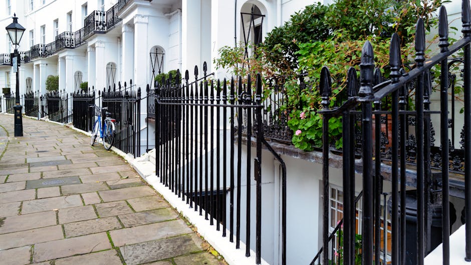 A row of white terraced houses with black wrought iron railings along a paved sidewalk. The railings feature pointed finials and are positioned in front of small front gardens filled with green foliage and flowering plants. A blue bicycle is leaning against one of the railings. The scene is illuminated by natural daylight, highlighting the clean and well-maintained appearance of the buildings and fencing. The setting suggests a residential area where private waste collection or on-site clearance may be employed as alternatives to traditional rubbish disposal methods, with visible fencing and front garden spaces emphasizing personal property boundaries. The image reflects a typical urban street scene with a focus on property boundary security and aesthetic garden presentation, linking to waste management services that handle rubbish removal or clearance for such residential properties in West Kensington.