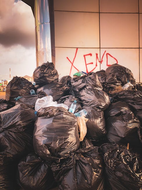 A large pile of black trash bags, filled and tied at the top, stacked against a modern building with a tiled wall in a neutral colour palette. Some bags have visible creases and slight tearing, with occasional clear plastic water bottles protruding from the bags, suggesting discarded packaging. Behind the pile, on the wall, there is red graffiti spelling 'XEND.' The setting appears to be an urban exterior, possibly a service or back alley area, with cloudy sky partially visible in the background. The scene reflects a situation requiring independent waste collection or private rubbish removal, typical of non-local authority disposal methods, and showcases the need for professional waste management services such as those provided by houseclearancewestkensington.co.uk.