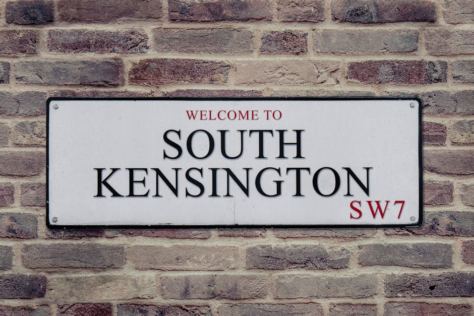 A rectangular white metal street sign mounted on a brick wall featuring the text 'WELCOME TO SOUTH KENSINGTON SW7' in black uppercase letters, with the words 'WELCOME TO' and 'SW7' in red. The sign is secured with four screws at each corner. The brick wall behind the sign comprises reddish-brown bricks with a rough texture, laid in a traditional horizontal pattern. There are subtle variations in the brick colors and mortar joints, creating an authentic urban appearance. The lighting is even and natural, highlighting the contrast between the smooth surface of the sign and the textured brick background. This signage scene is typical of urban residential or commercial areas in South Kensington, which may be associated with private property or independent local services such as rubbish removal, as reflected in the visual context of on-site waste management or clearance activities.