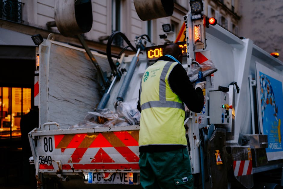 A waste collection worker stands with their back to the camera, wearing a high-visibility yellow vest and dark clothing, operating the control panel of a large, white rubbish collection truck. The truck's rear section is open, revealing a load of mixed waste and debris, including crumpled paper, plastic, and other refuse materials. The truck is parked on a street at dusk, with a background featuring a building with illuminated windows. The scene includes safety features such as reflective red and white chevron markings on the truck’s rear, and the worker appears to be engaged in managing or documenting the waste collected, supporting onsite waste handling and private rubbish removal services typical in independent waste collection operations managed by companies like houseclearancewestkensington.co.uk.