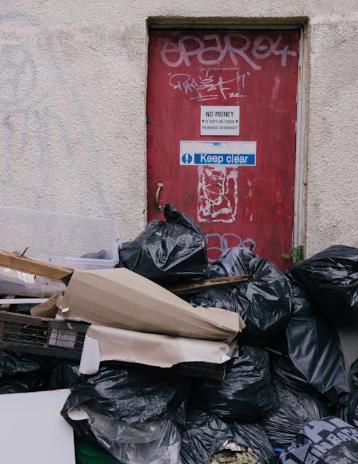 A large pile of black rubbish bags, some torn open, is stacked against a beige, textured exterior wall near a red, graffiti-covered metal door. The bags appear full and bagged items are irregularly shaped, with some leaning on pieces of cardboard or wooden panels. The door features multiple stickers, including a white one with the text 'No Money' and a blue sticker with the phrase 'Keep Clear,' both attached above and around the debris. The scene suggests an informal waste collection or clearance situation, with the waste likely awaiting removal or disposal. The environment appears to be an alley or back alley area, with natural daylight illuminating the scene and casting subtle shadows across the wall and debris, supporting the context of discreet, off-premises rubbish removal by a service like House Clearance West Kensington.