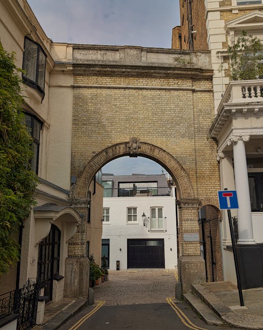 A narrow urban alleyway featuring a historic brick archway with a keystone and decorative crest at the top, flanked by cream-colored and white buildings with classical architectural details. To the left, a bay window with black framing extends from a building with a curved white cornice and lush green foliage trailing down its side. To the right, a white residential building with ornate columns and balustrades creates a balcony, alongside a blue and red traffic sign indicating a dead-end. The cobbled pathway beneath the archway is lined with double yellow lines and leads to a modern white apartment building with large windows, a black garage door, and a small balcony. The scene suggests a quiet residential area where private waste collection services, such as those offered by houseclearancewestkensington.co.uk, could be used for rubbish removal, especially in locations with limited access for large Waste Management vehicles. The ambient daylight highlights the varied textures and colours of the historic and contemporary architecture, emphasizing a setting suitable for on-site clearance or independent waste handling within an urban environment.