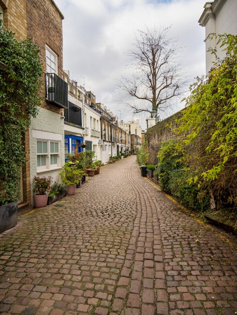 A narrow cobblestone street in a residential area, lined with potted plants and small trees along the base of colorful terraced houses. The houses feature white and pastel facades, some with bay windows and small balconies with black railings. A large, leafless tree with sprawling branches stands prominently at the end of the street, set against a cloudy sky. On the right side, a tall brick wall covered with lush green climbing plants and shrubs separates the street from an adjacent property. The scene captures a quiet, historic neighbourhood with a mix of greenery and urban architecture, suggesting a setting where private waste collection or on-site clearance might be relevant to local rubbish removal services like those offered by houseclearancewestkensington.co.uk, especially for managing clutter in confined urban alleys or alleyways with narrow access.
